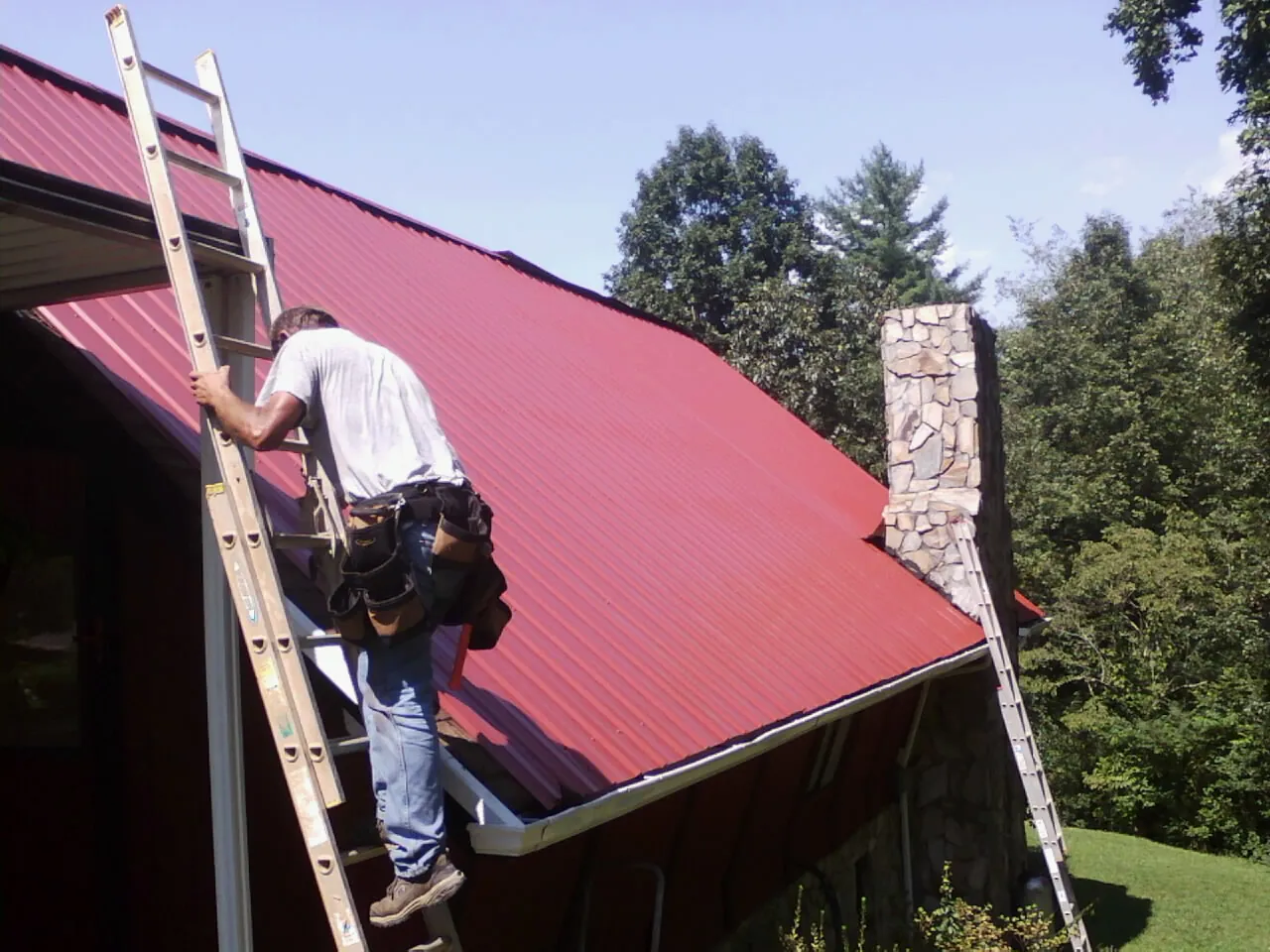 Roofer on a ladder beside a red metal roof and stone chimney — Soffit & Fascia Repair by Roofing Brothers in Southfield